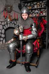 A young knight in medieval armor with a sword in his hand sits in a rocking chair by the fireplace.