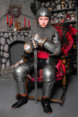 A young knight in medieval armor with a sword in his hand sits in a rocking chair by the fireplace.