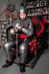 A young knight in medieval armor with a sword in his hand sits in a rocking chair by the fireplace.