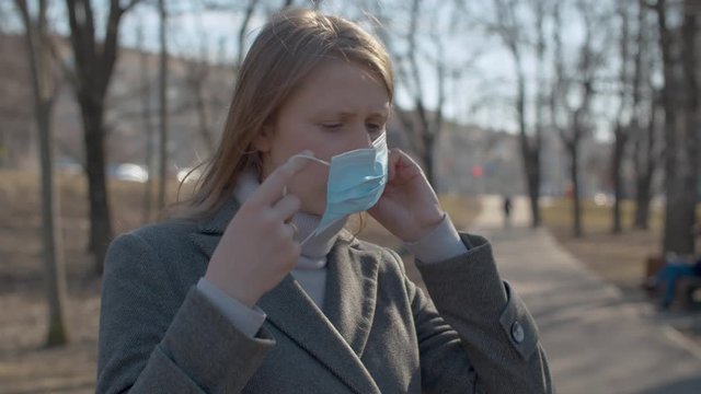 Close up portrait of a young pretty woman putting on a protective mask outdoors in the city park. Sunny sping day. Protect yourself and your loved ones. Covid-19 pandemic