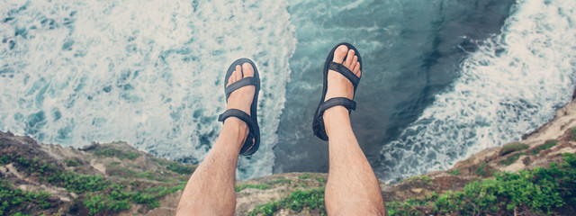 Young brave man sitting on a high cliff above stormy ocean (intentional vintage color, POV view). Wide screen panoramic