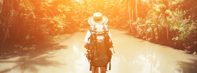 Traveling woman with backpack and straw hat looking at tropical river at sunny day. Wide screen
