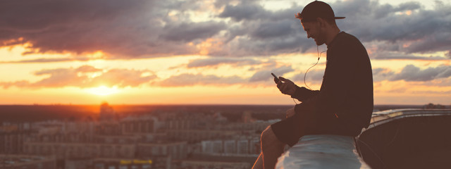 Young brave man sitting on the edge of the roof with smartphone. Wide screen panoramic