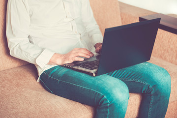 Naklejka premium young man in white shirt and blue jeans working on laptop, writes in notebook in home office sitting on cozy beige sofa Stay at home, online work, distance learning concept