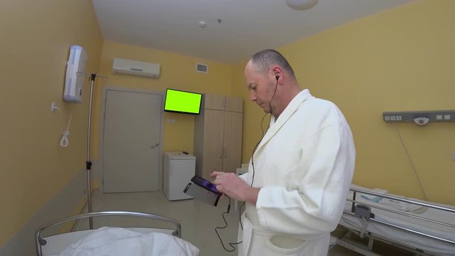 Male patient in hospital gown searches for news on smartphone with headphones. Chromakey green in the background in hospital boxing adult man in a white hospital robe with a smartphone in his hands.
