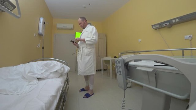 Patient in hospital room tries to distract himself by listening to music on his smartphone through headphones. The ruffled bed medical functional furniture white hospital gown and state sneakers good.