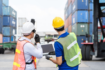 Foreman manual worker at commercial dock, wearing yellow, white safety helmet, holding radio communication, laptop, notebook for control loading container box from cargo freight ship for import export