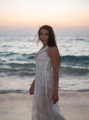 Fototapeta na wymiar Romantic portrait of beautiful brunette woman in white lace dress standing at the beach over sea and sunset sky background