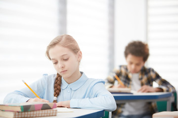 Portrait of cute blonde girl writing test while sitting at desk in school classroom, copy space