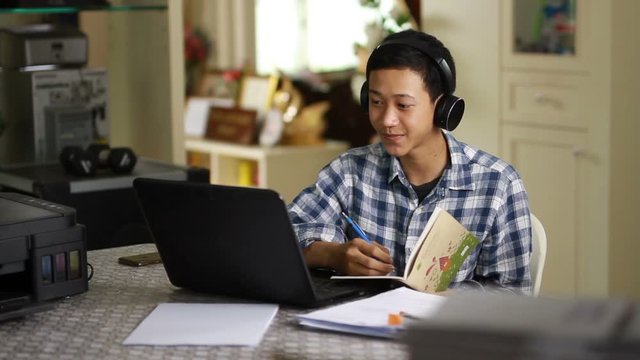 Teenager of Asian man wearing an earphone while learning by using a laptop at home. online course and working at home concept.