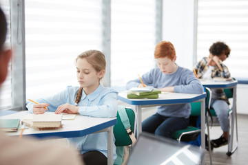 Side view at multi-ethnic group of children sitting in row at desk in school classroom and writing or taking test, copy space - obrazy, fototapety, plakaty