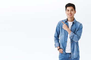 Portrait of enthusiastic handsome taiwanese man in denim jacket, pointing finger upper left corner, give recommendation, suggest visit page, click link to online shop, smiling camera