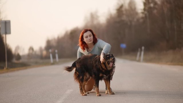 Young redhead woman plays with pariah dog during sunset in middle of road. Slow motion