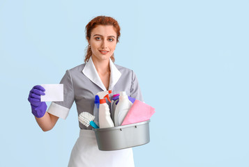 Beautiful young chambermaid with business card and cleaning supplies on color background