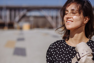 Obraz premium Beautiful girl model stands on the background of a bridge in the city and enjoys the warm spring weather. Close-up. Smiling with eyes closed.