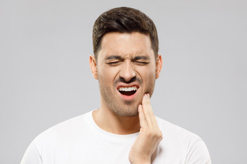 Fototapeta premium Toothache concept. Young man touching his chin and closing eyes with expression of suffer from tooth ache, isolated on gray background