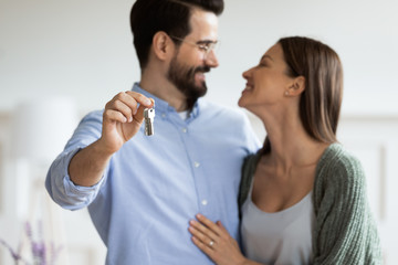 Focus on smiling bearded man in eyewear showing keys to camera, cuddling attractive wife, head shot. Happy family couple homeowners celebrating moving into new apartment, real estate concept.
