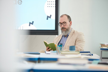 Portrait of senior bearded professor sitting at desk in school classroom and reading book, copy space