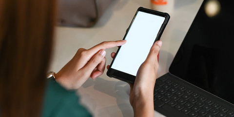 Cropped image of hands holding a cropped black smartphone with white blank screen over the working desk as background.