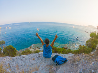 Young woman with backpack sitting on a cliff looking at the ocean spreading arms. Ponza Island coast, Italy.