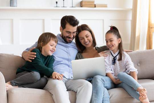 Smiling parents cuddling little kids, looking at laptop screen. Happy family couple watching funny movies or cartoons, having fun together in living room, relaxing on sofa, enjoying weekend time.