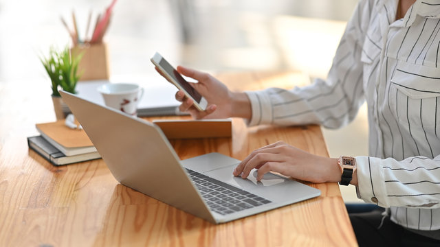 Cropped image of young creative woman holding a smartphone in hand while typing on computer laptop that putting on wooden working desk over living room as background. Remote working concept.