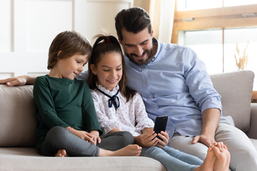 Smiling adorable little schoolgirl sitting on couch, showing funny video on smartphone to younger brother and smiling father in living room. Addicted to tech dad enjoying leisure time with children.