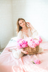 Cute tender young girl with blond hair in white clothes and lace underwear with a basket of pink peonies on the bed in the studio. Spring and flowers. Beauty and Fashion