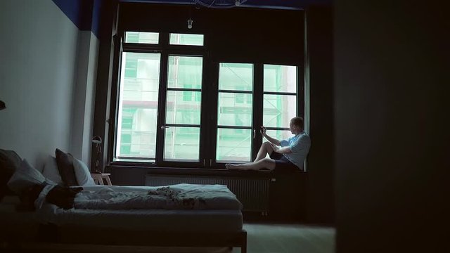 silhouette of a man sitting on a windowsill near a window during a quarantine or emergency. in a bedroom apartment with large windows and a bed.  dark room