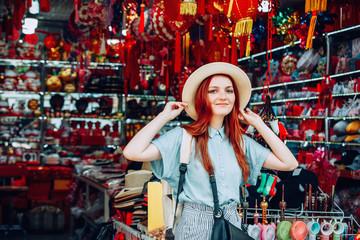 happy young caucasian red hair woman wearing blue shirt and black purse tries on a hat while shopping at store sells wicker hats in Asian touristic market in China. travel shopping lifestyle concept