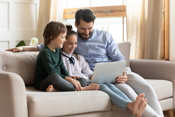Fototapeta premium Smiling young dad resting on couch with cute children, showing educational applications on computer. Happy man enjoying spending free leisure time with kids son daughter, watching movie at home.