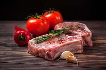  Pork steak with spices, vegetables and rosemary. Juicy and fresh raw meat, tomatoes, paprika, henna, and peppercorns on a wooden tabletop