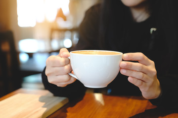 A woman hands holding hot cup of coffee or tea in morning sunlight at the cafe.