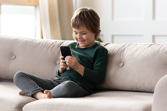 Smiling little preschool child boy playing online game, watching video on cellphone, entertaining alone in living room. Happy small kid using funny mobile apps, enjoying free leisure time at home.