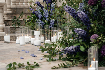 Burning white candles in glass vases on the floor near the greenery and flowers. Details of the wedding ceremony.
