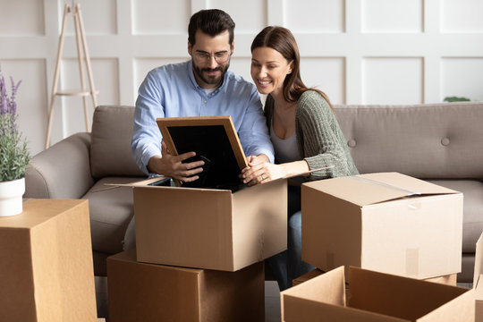 Full length happy young family couple unpacking belongings, looking at family photo in frame. Smiling spouses unboxing decorations, sitting together on comfortable couch between cardboard boxes.