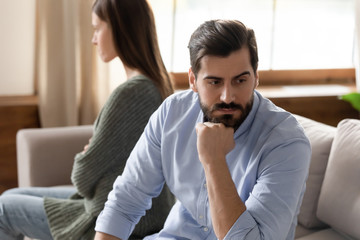 Focus on stressed thoughtful young man sitting separate from offended wife on couch at home. Young...