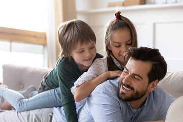 Playful small kids siblings lying on fathers back, having fun together on comfortable sofa at home. Smiling young dad holding on back cute children son daughter, playing on couch in living room.