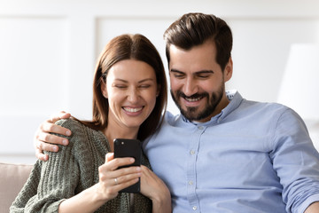 Head shot close up laughing young woman sitting on couch with joyful boyfriend husband, looking at smartphone screen. Happy married family couple watching funny video, making selfie on mobile.