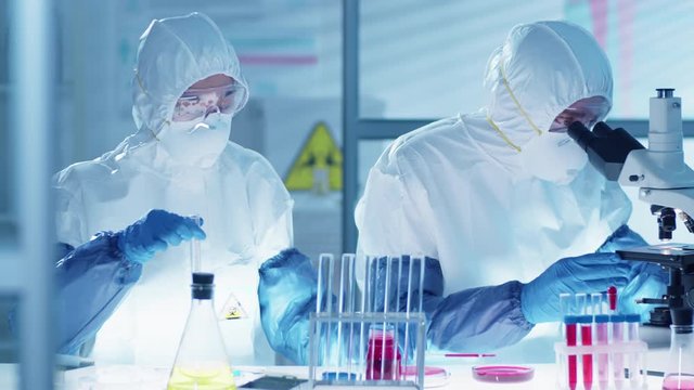 Two lab workers in protective suits, glasses, masks and gloves conducting research on antiviral treatment together: female scientist pouring blood into test tube and man looking through microscope