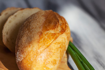 An appetizing bun with a golden crust is cut into slices, lying on a small kitchen board next to a fresh green onion in the rays of sunlight. Healthy breakfast.