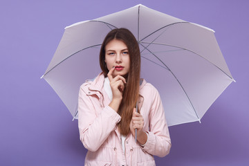 Horizontal photo of thoughtful attentive attractive female holding umbrella, putting one finger on cheeck, having pensive facial expression, wearing casual clothes. Weather forecast concept.