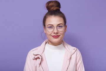 Close up portrait of delighted charming model looking directly at camera, standing isolated over lilac background in studio, wearing eyeglasses, having bun, wearing casual clothes. Youth concept.