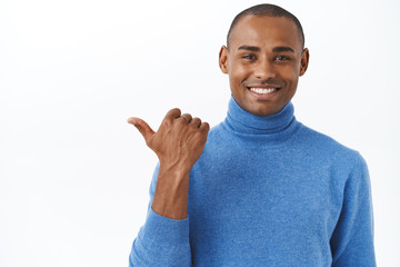 Check out my new car, its outside. Portrait of pleased, proud smiling african american male pointing thumb left and grinning satisfied, showing banner, promo of company or service, white background