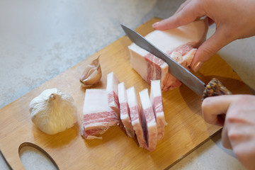 Fototapeta na wymiar Horizontal indoor picture of unknown person holding knife, cutting lard into slices, preparing for lunch. Salo and garlic being on wooden cutting board, traditional food. Eating habits concept.