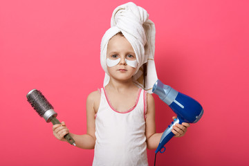 Portrait of cute good looking little child holding round hair brush and hair dryer in both hands, having patches under eyes, wearing pajama, having towel on head, taking care, hygiene and cleaning.