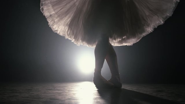 Close up of ballet dancer as she practices exercises on dark stage or studio. Woman's feet in pointe shoes. Ballerina shows classic ballet pas. Slow motion. Flare shot.