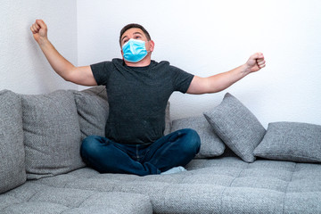 A bored cheerful man in quarantine in a medical mask is yawning and stretchingsitting on a sofa and is bored. Self-isolation coronavirus quarantine at home. Pandemic COVID19
