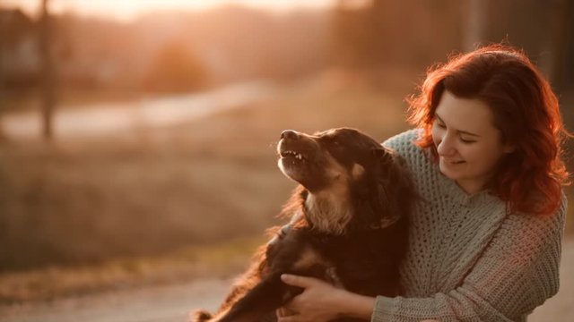 Cute redhead woman shows tongue and hugs and plays with cur dog during sunset on blurry warm background. Slow motion