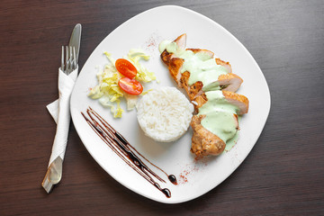 Chicken fillet with sauce is served with rice and vegetables on a white plate and a dark table. Top view.